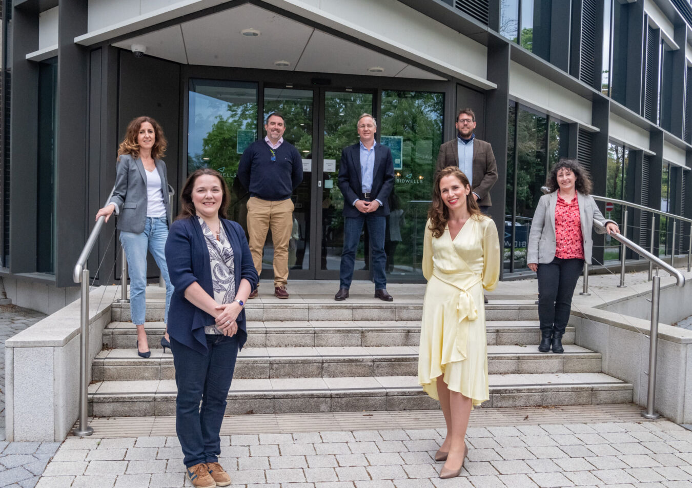 Seven people standing on steps outside a modern building with glass doors, posing for a group photo. The building has "Bidwells" signage on the door.