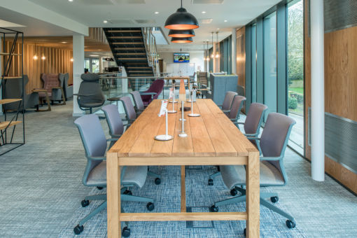 Contemporary open-plan workspace with a long wooden communal table, ergonomic chairs, individual desk lamps, and large floor-to-ceiling windows providing natural light