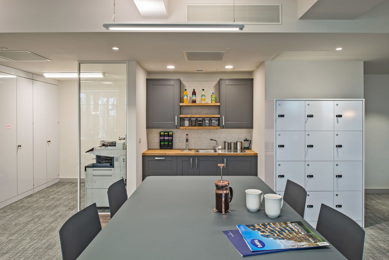 Modern office kitchen with gray cabinets, a countertop with coffee equipment, lockers on the right, and a table with a brochure and coffee set