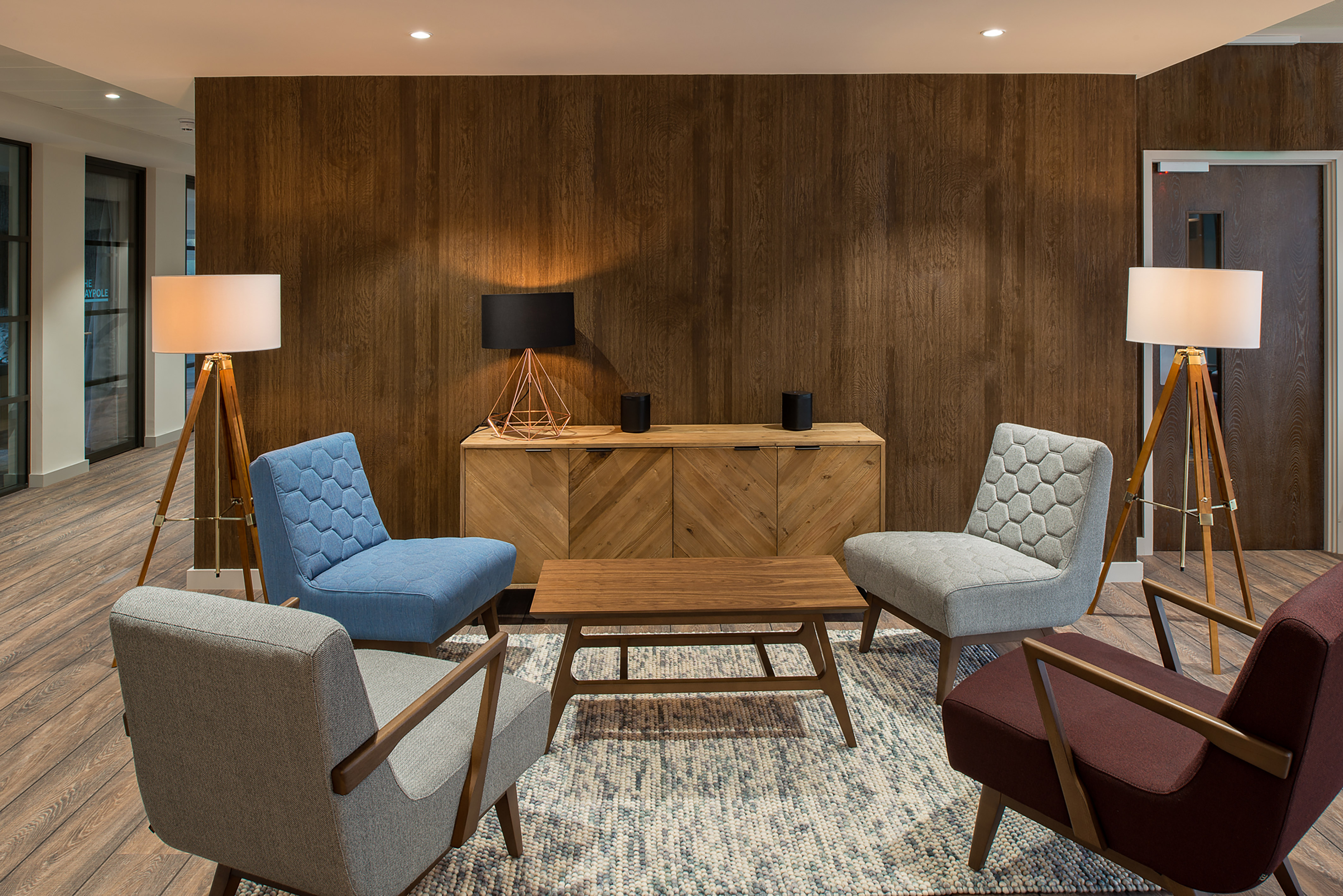 Stylish modern lounge with four mid-century armchairs around a wooden coffee table. Warm lighting from tall lamps, wood panel walls, and cozy ambiance