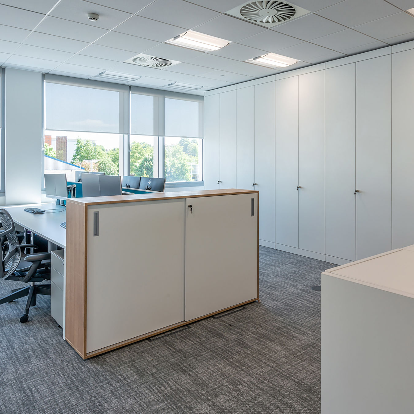 Bright, modern office with gray carpet, white walls, large windows with blinds, and sleek workstations featuring computers and ergonomic chairs