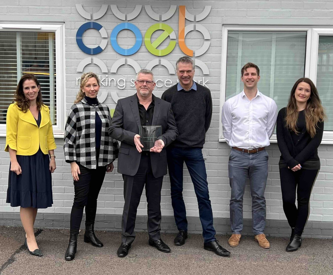 Seven individuals stand smiling in an outdoor setting; the central person holds a glass award. Behind them, a wall displays the text "coel making space work."