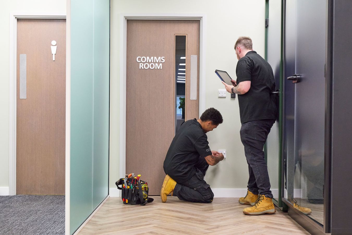 Two technicians work near a wall socket; one kneels adjusting it, the other holds a tablet. A tool bag is on the floor beside them, next to a "COMMS ROOM" door.