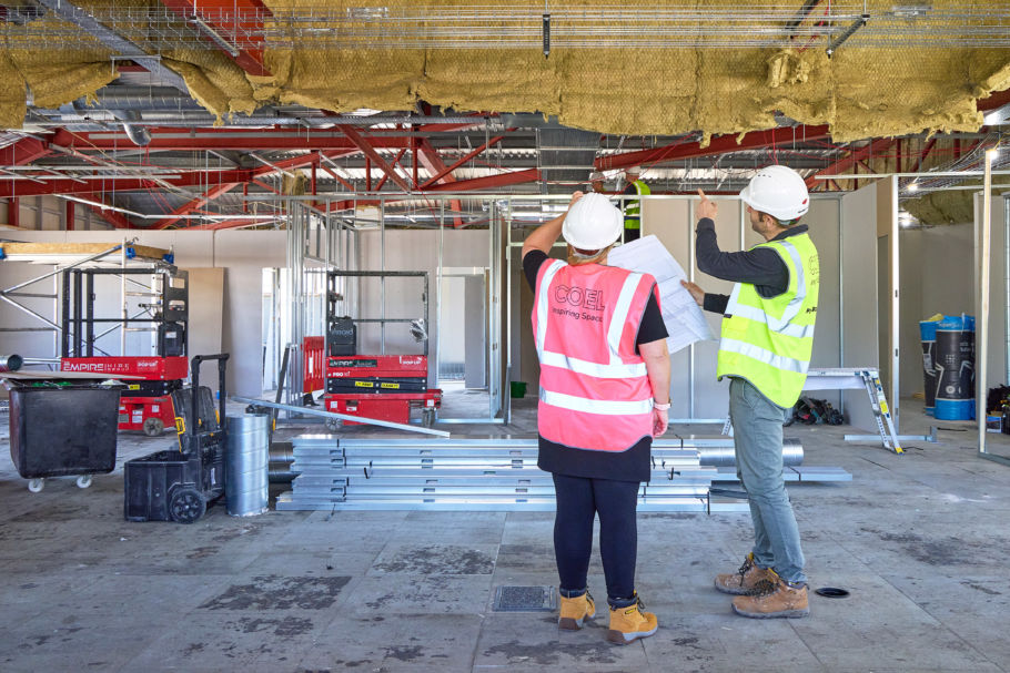 Two construction workers in safety vests and helmets review blueprints in a partially built office with exposed ceilings and scattered metal beams