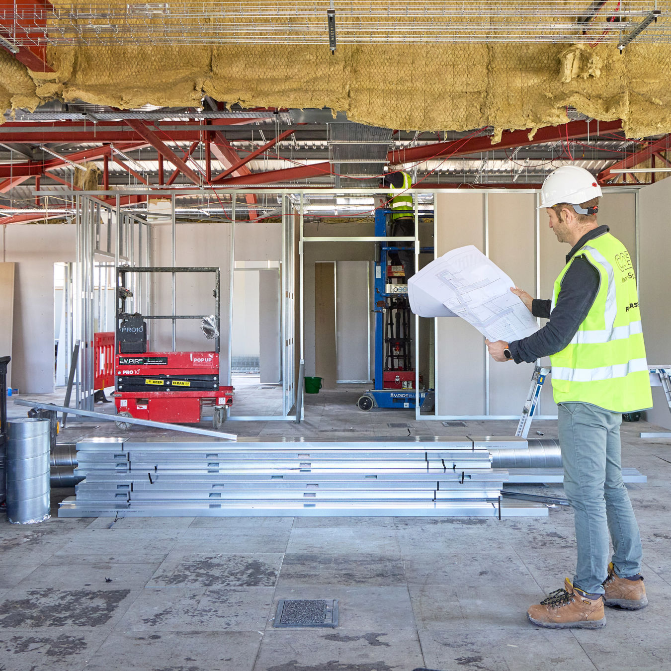 A construction worker in a hard hat and safety vest examines blueprints in a half-built room with exposed insulation, metal beams, and scaffolding.