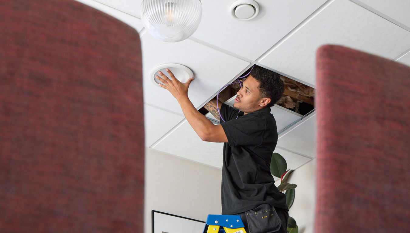 A person on a blue stepladder installs a ceiling fixture in an office with white ceiling tiles and burgundy dividers in the foreground.