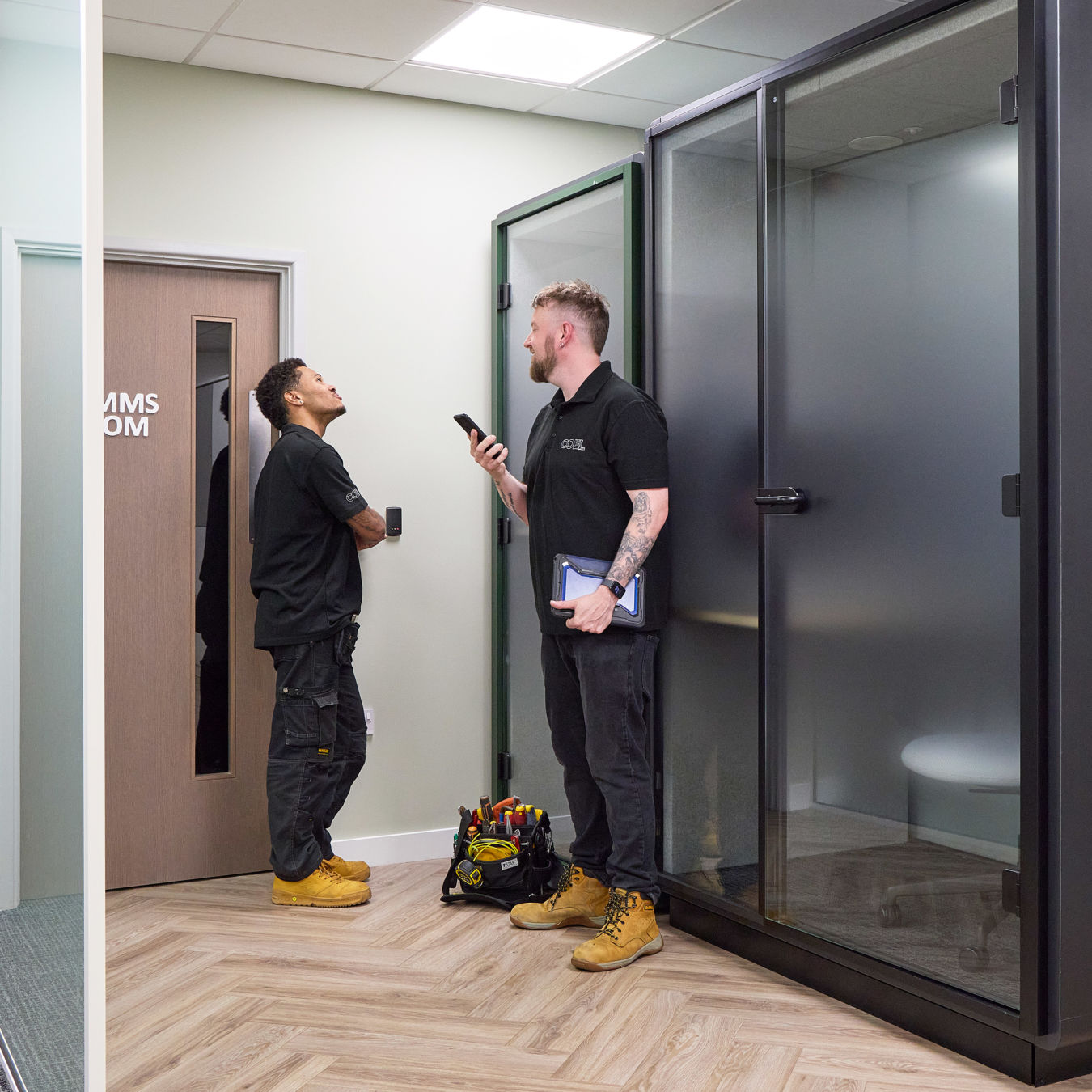 Two workers in black shirts converse in an office hallway. One holds a phone, the other leans against a door labeled "COMMS ROOM." A tool bag sits on the floor.