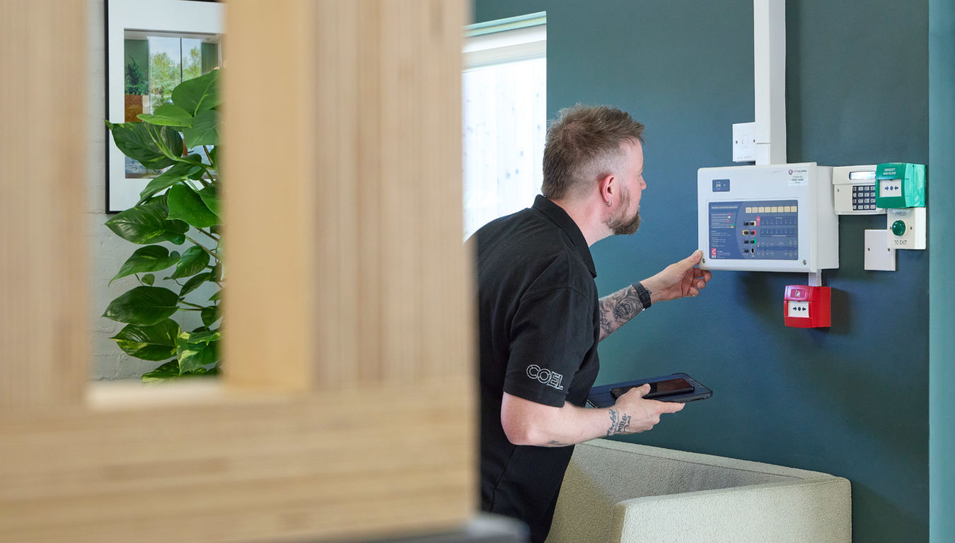A man interacts with a wall-mounted security panel, holding a tablet in a modern office with green walls and plants. Text visible on clothing: "COEL".