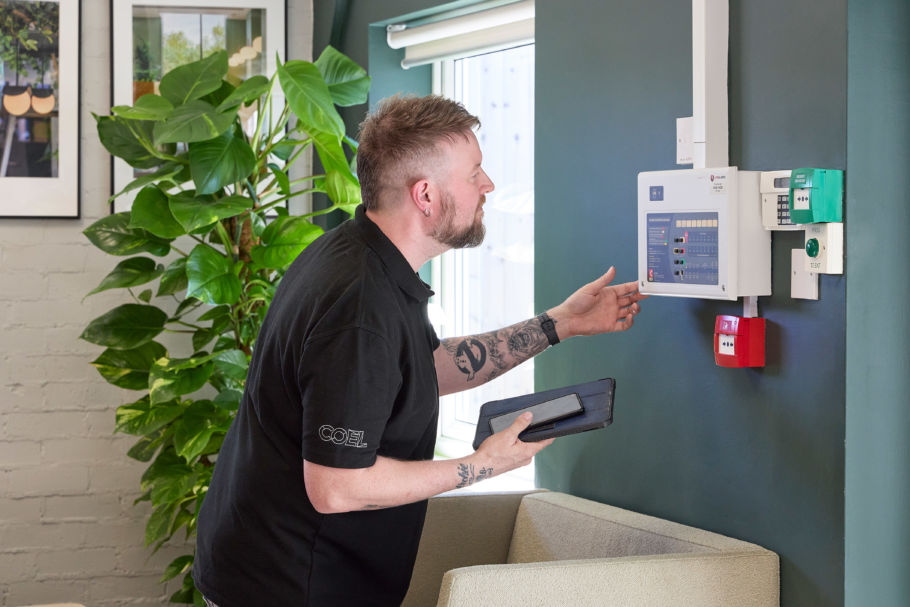 A man examines a control panel mounted on a green wall, holding a tablet, with a large plant nearby and sunlight streaming through a window. The shirt text reads "COEL."