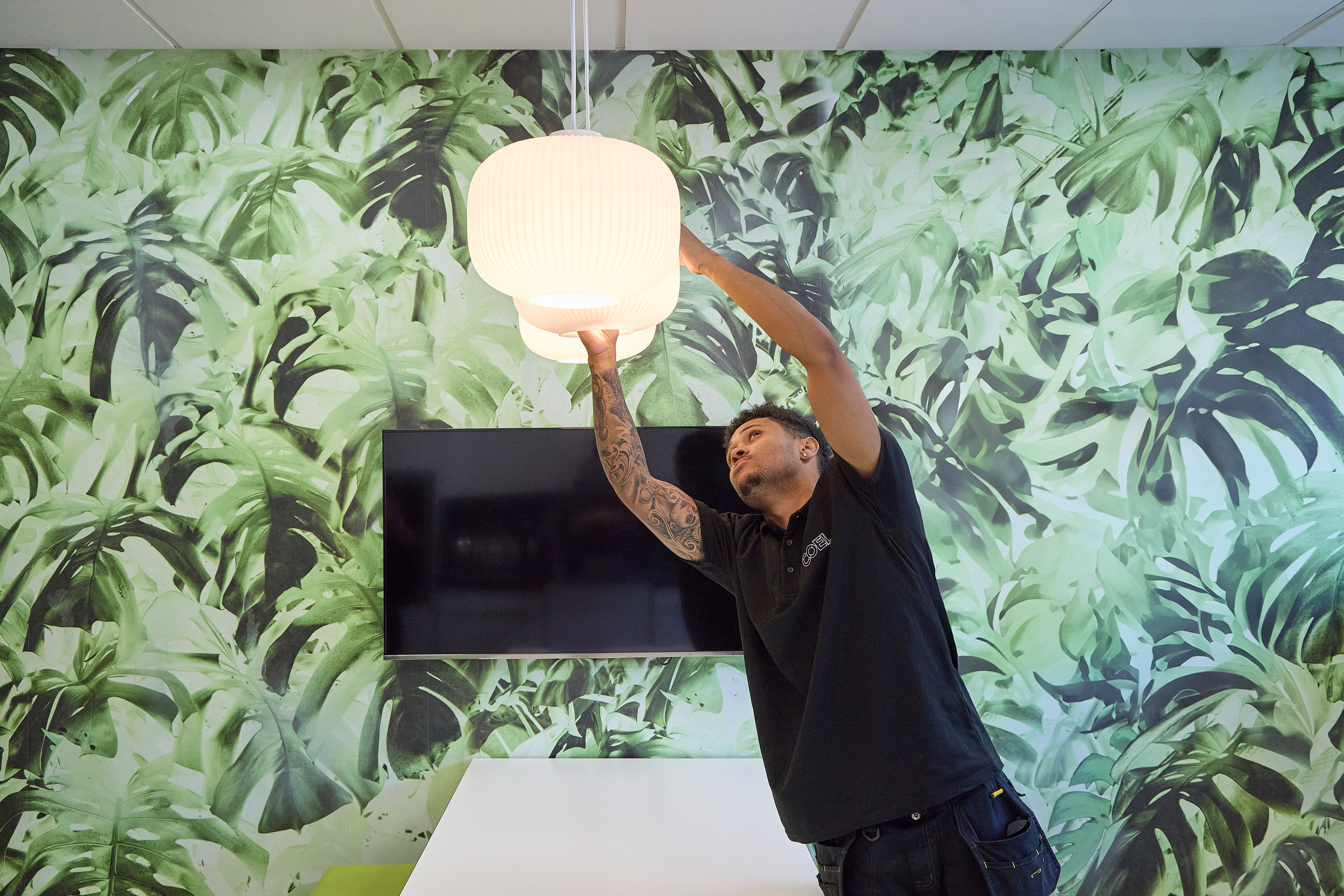 A person adjusts a white pendant light in a room with green leafy wallpaper and a mounted television screen.