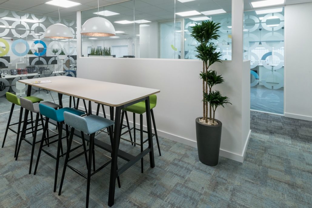 A modern office space features a high table with multicolored stools, illuminated by pendant lights, next to a partition wall and decorative plant, with frosted glass and geometric patterns.