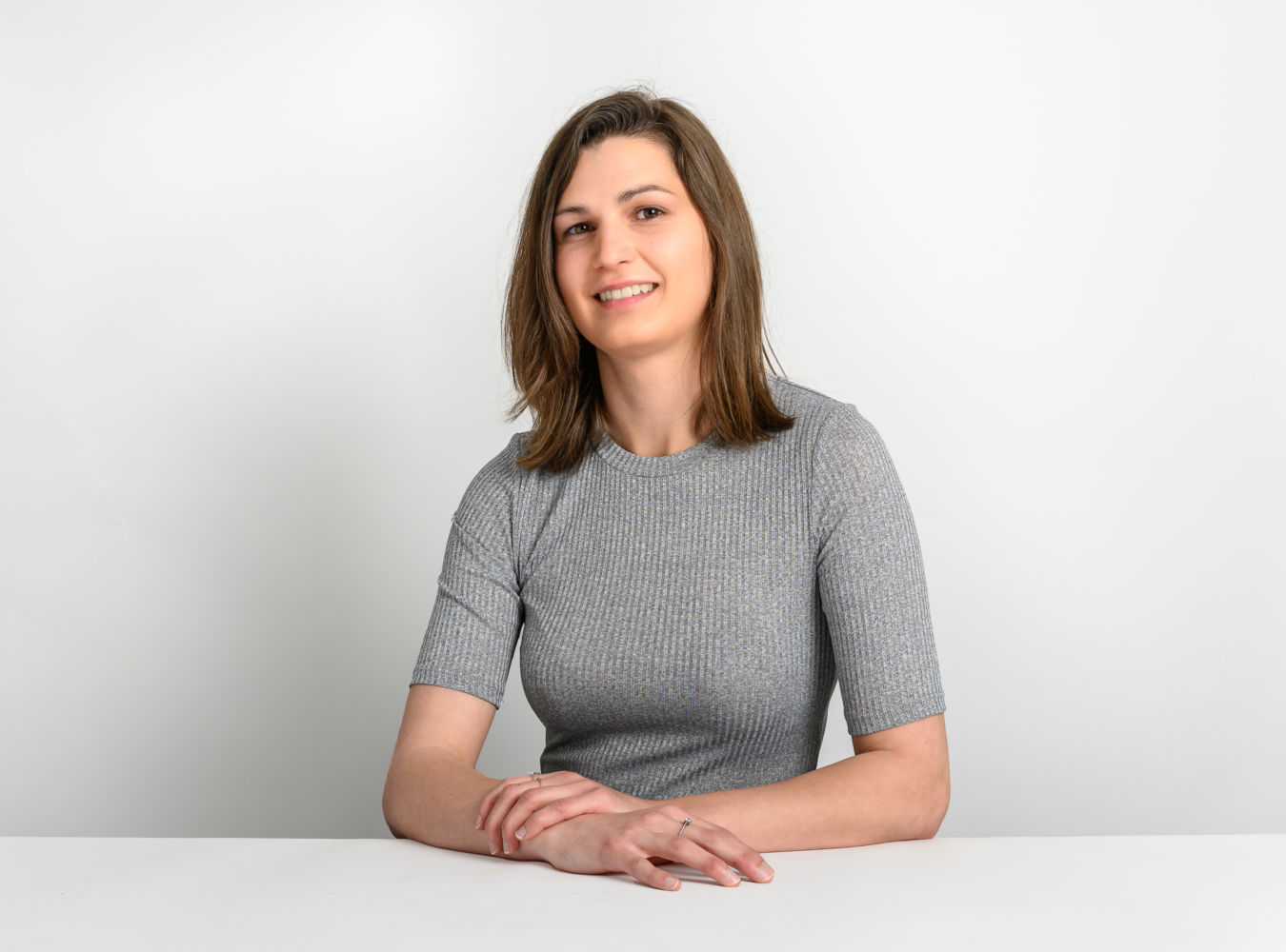 A person with shoulder-length brown hair smiles while seated at a white table in a gray ribbed shirt against a plain white background.