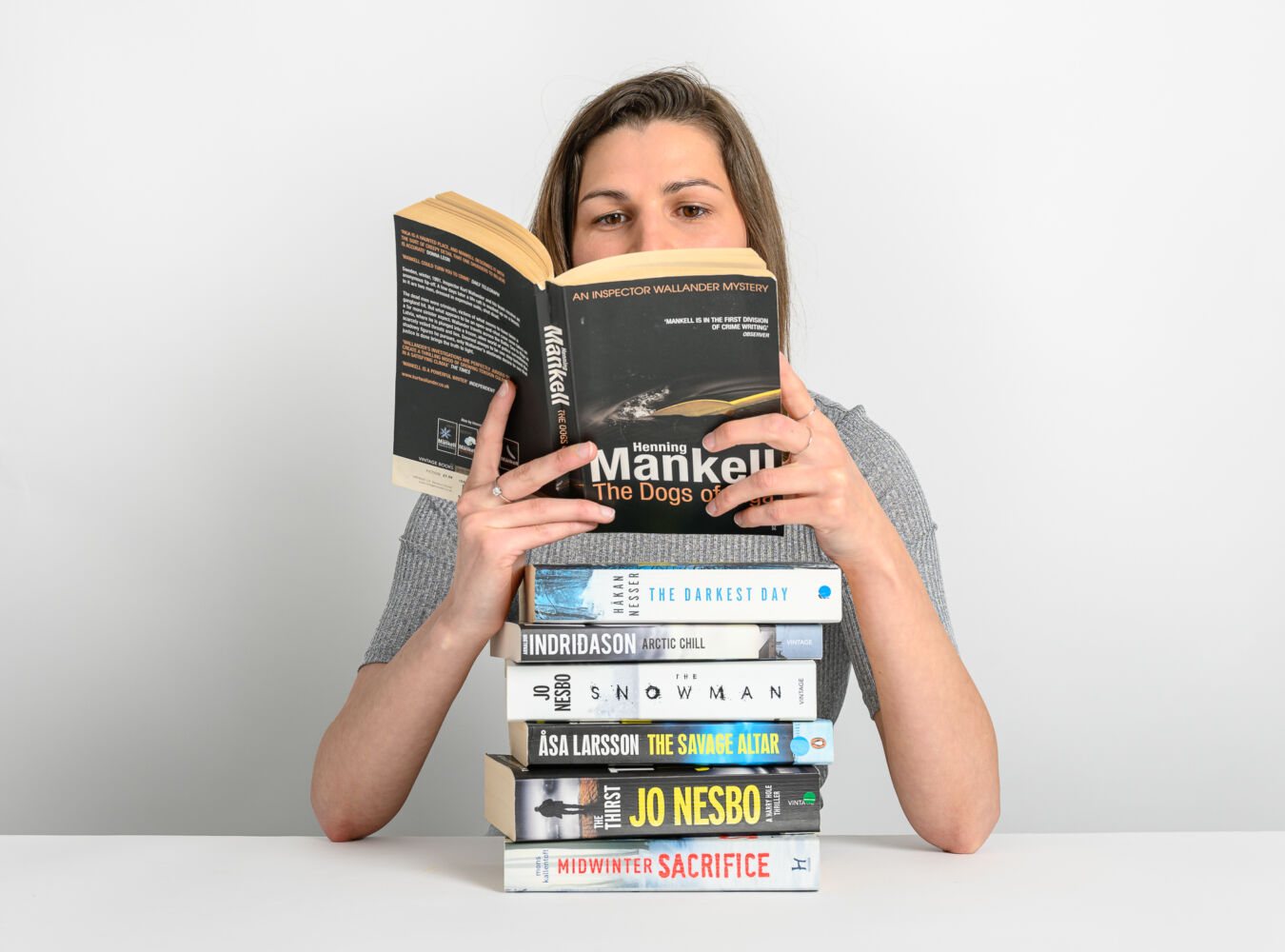 A woman reads "The Dogs of Riga" by Henning Mankell, holding it above a stack of crime novels titled "The Darkest Day," "Arctic Chill," "The Snowman," "The Savage Altar," "Thirst," and "Midwinter Sacrifice."