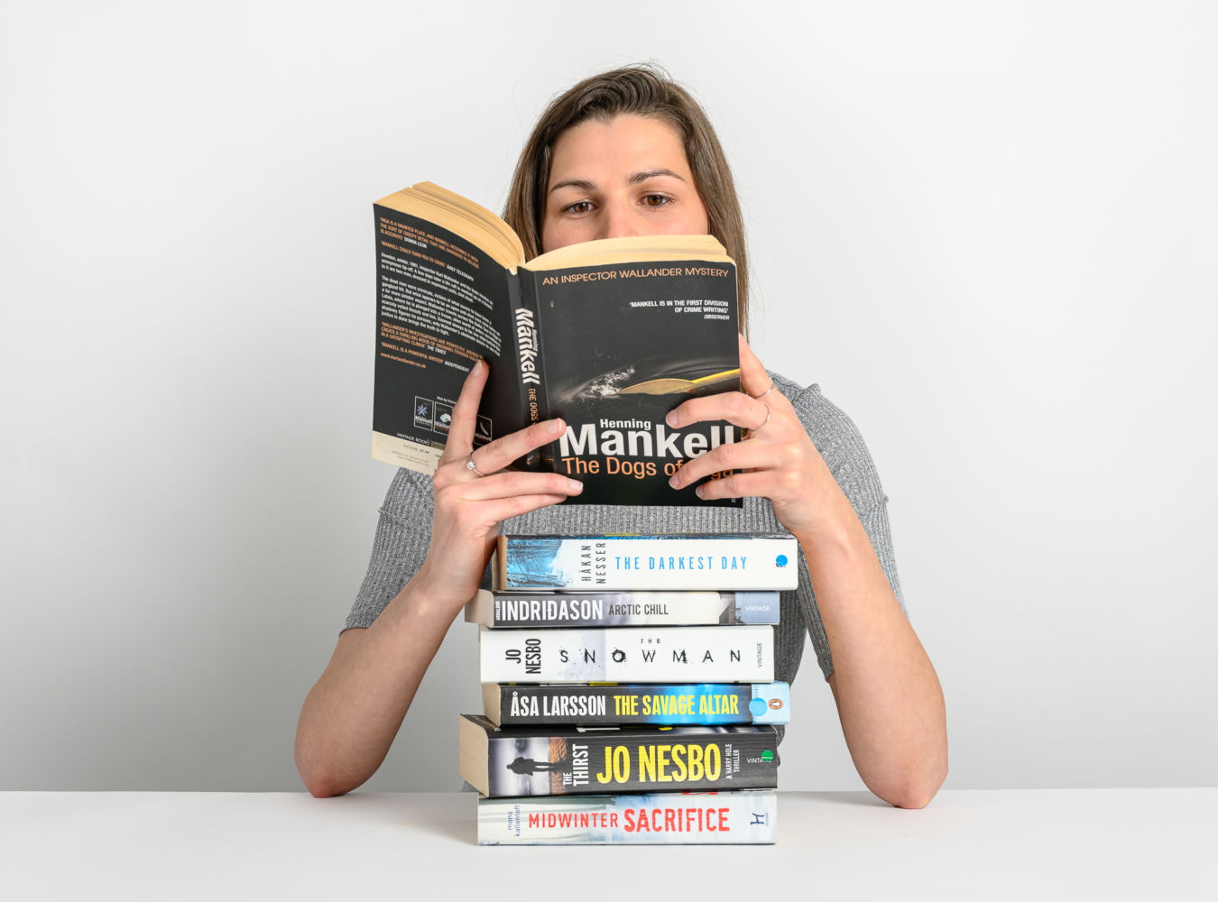 A person reads "Henning Mankell: The Dogs of Riga" over a stack of crime novels, titled "The Darkest Day," "Arctic Chill," "The Snowman," "The Savage Altar," "The Thirst," "Midwinter Sacrifice," on a white table.