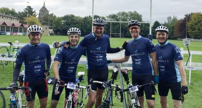 Five cyclists in blue "Bidwells" jerseys stand together, smiling with bicycles, in a grassy outdoor sports field. Each bike has a number tag, and a rugby goalpost is visible behind them.
