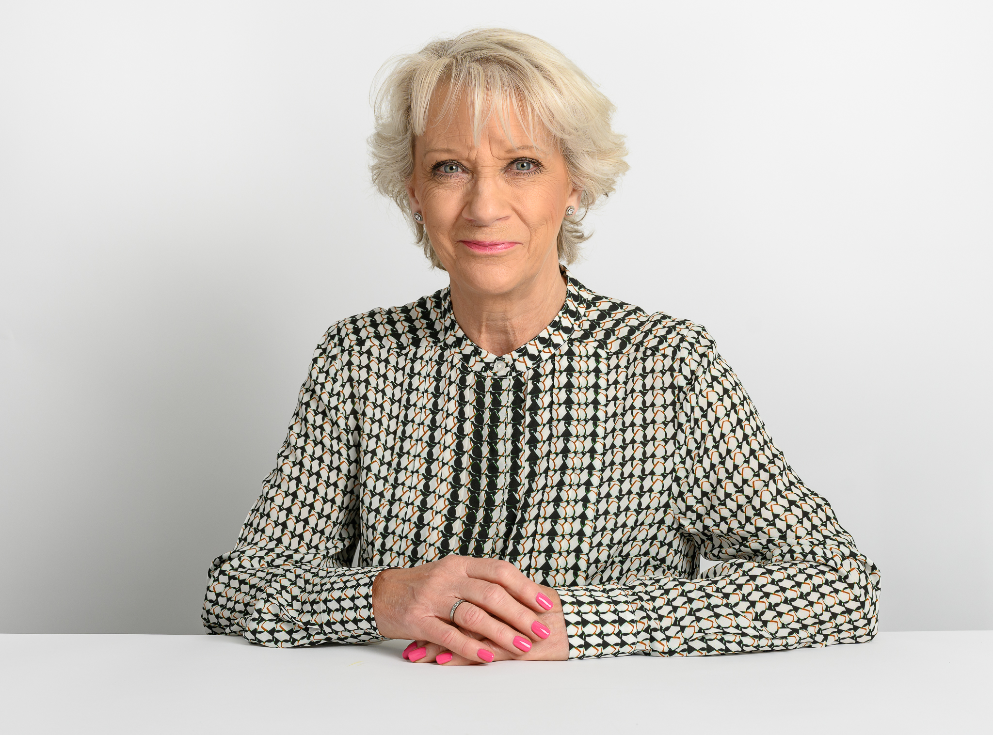 A woman sits with her hands clasped on a white table, wearing a patterned blouse and displaying pink-painted nails, against a plain white background.