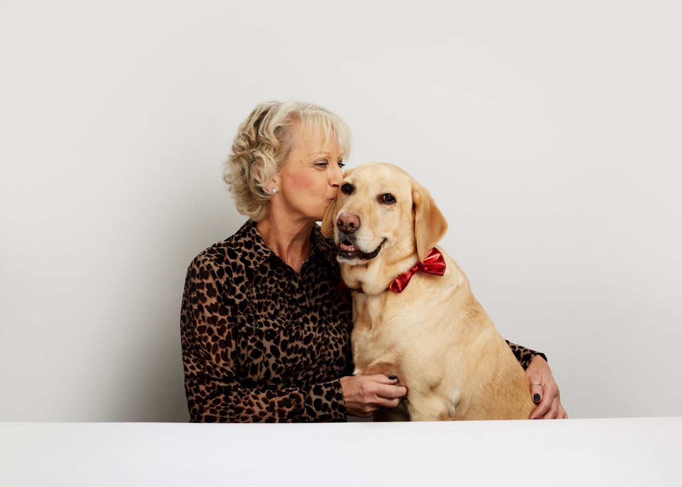 A woman in a leopard print top kisses a Labrador retriever wearing a red bow tie, sitting closely in a bright, minimalistic setting.