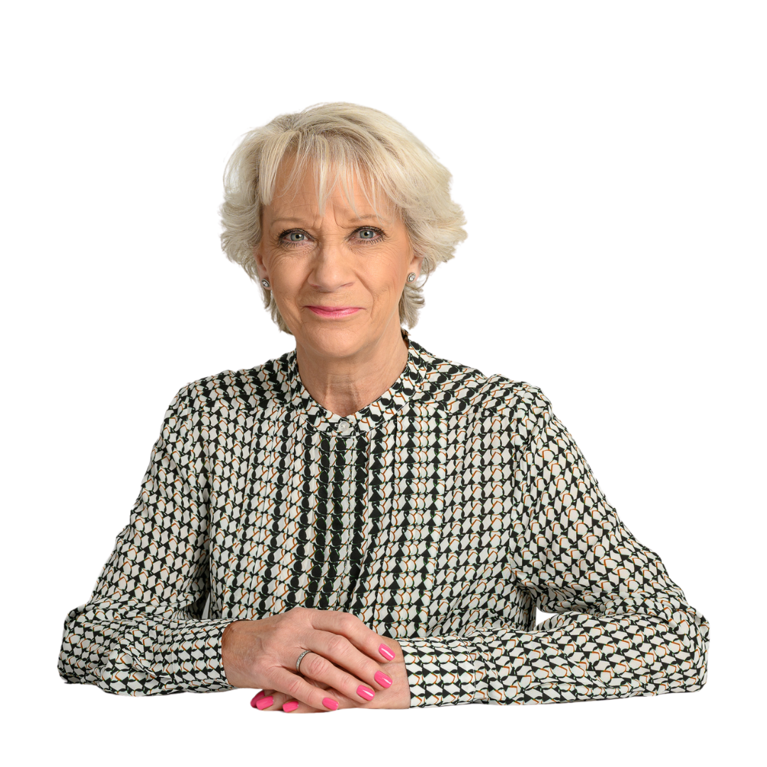 A woman with short, light hair and pink nails sits with her arms crossed, wearing a patterned blouse against a plain black background.