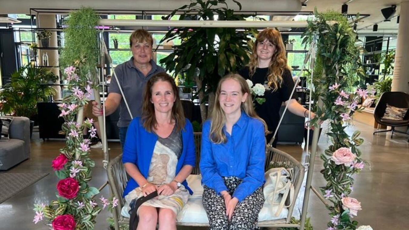 Four people gathered around a decorative floral swing in a modern indoor setting with large plants and stylish furniture.