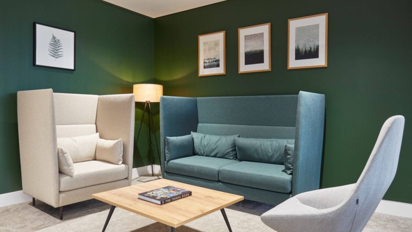 Modern lounge area features beige and teal armchairs around a wooden coffee table with "The Travel Book." Framed artwork decorates the green wall, and a floor lamp provides warm lighting.