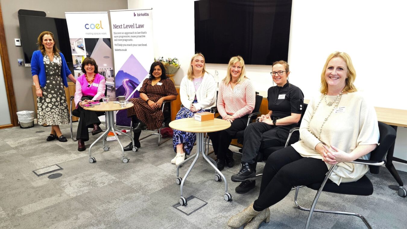 Seven women seated and smiling in a conference room, with two banners reading "coel making space work" and "Next Level Law birketts.co.uk" behind them. A table holds drinks and a box.