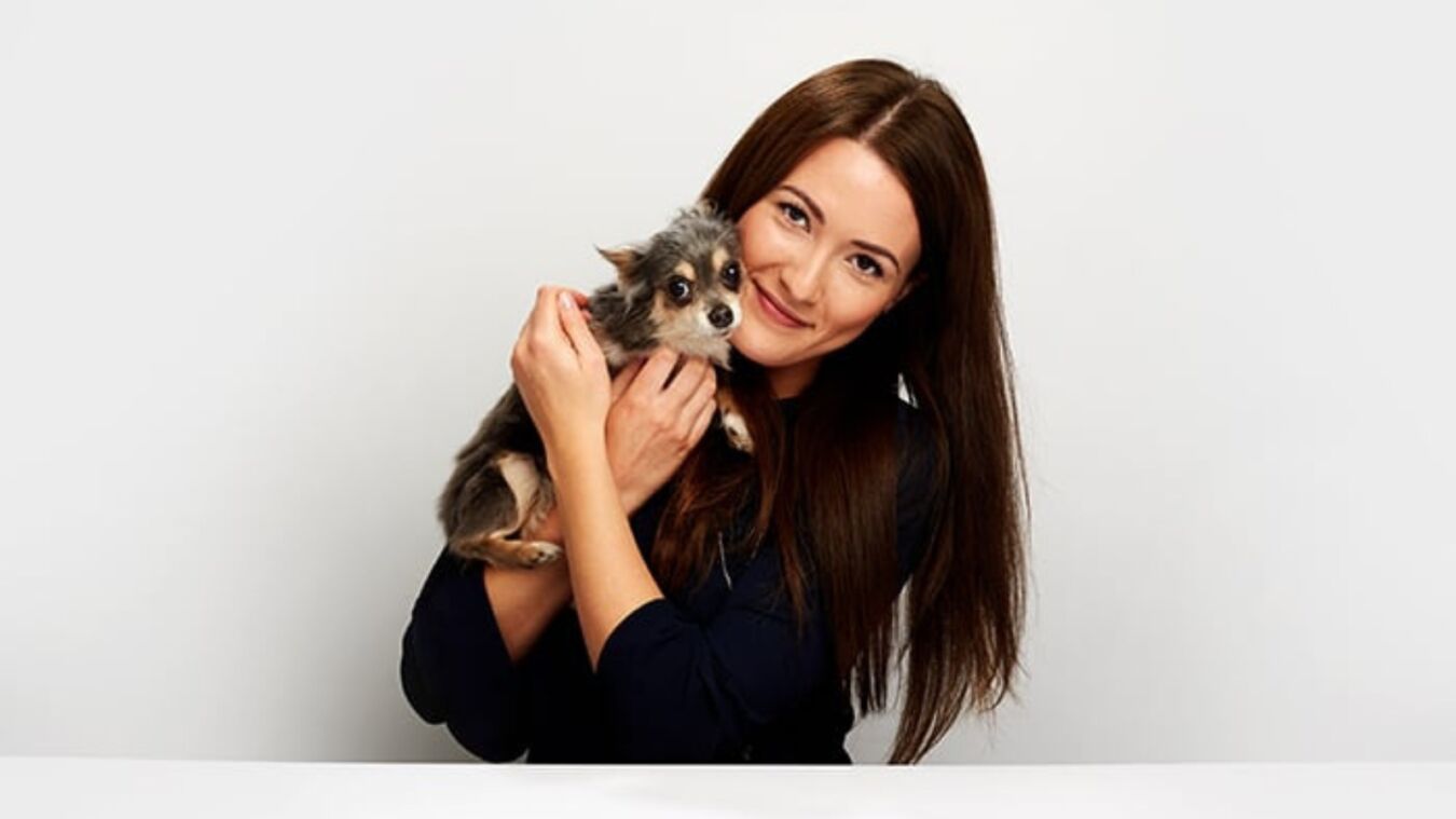 A woman with long brown hair holds a small dog closely, smiling against a plain white background.
