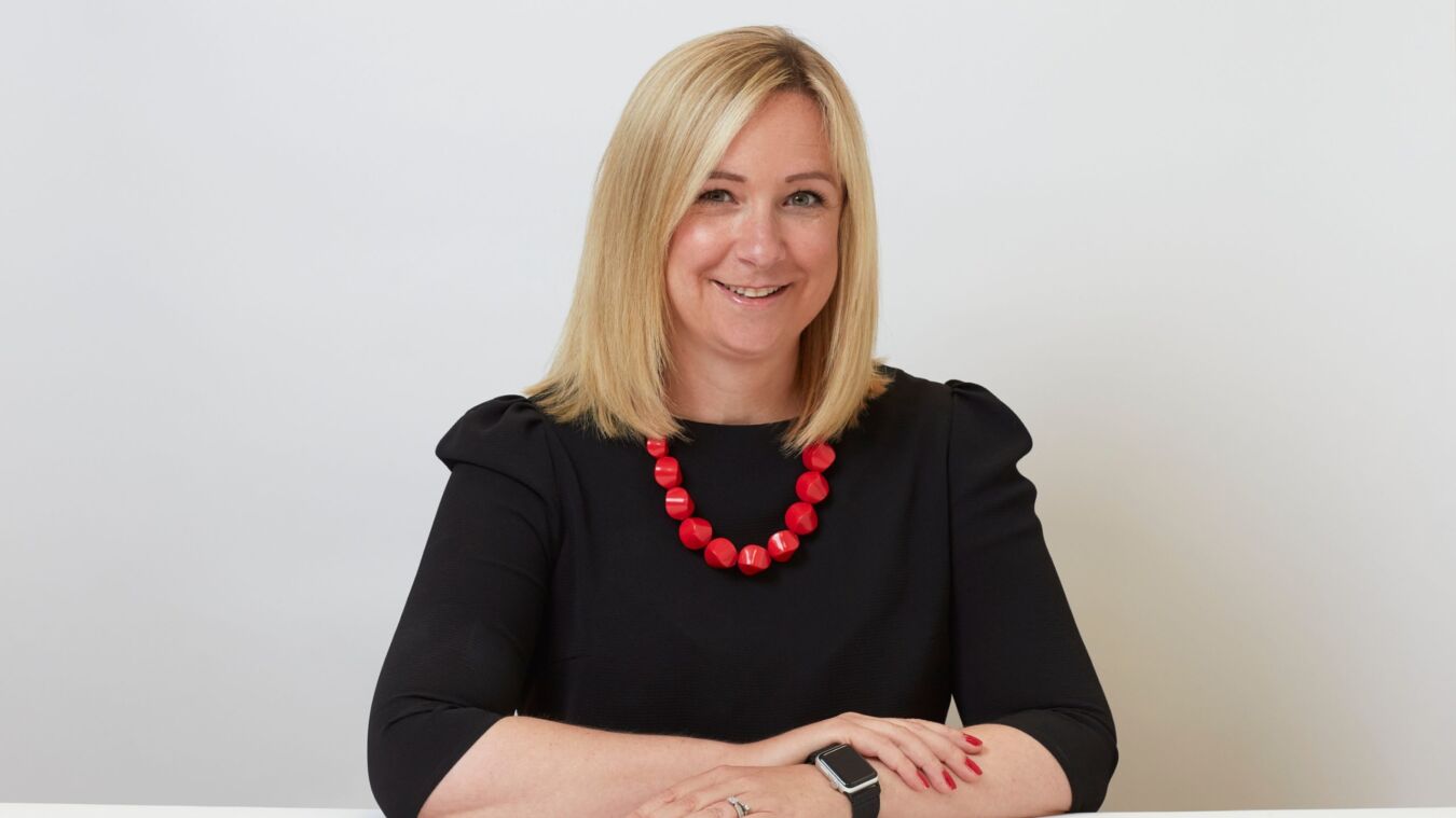 A person with blonde hair smiles, wearing a black top and red beaded necklace, sitting at a white table with a plain background.