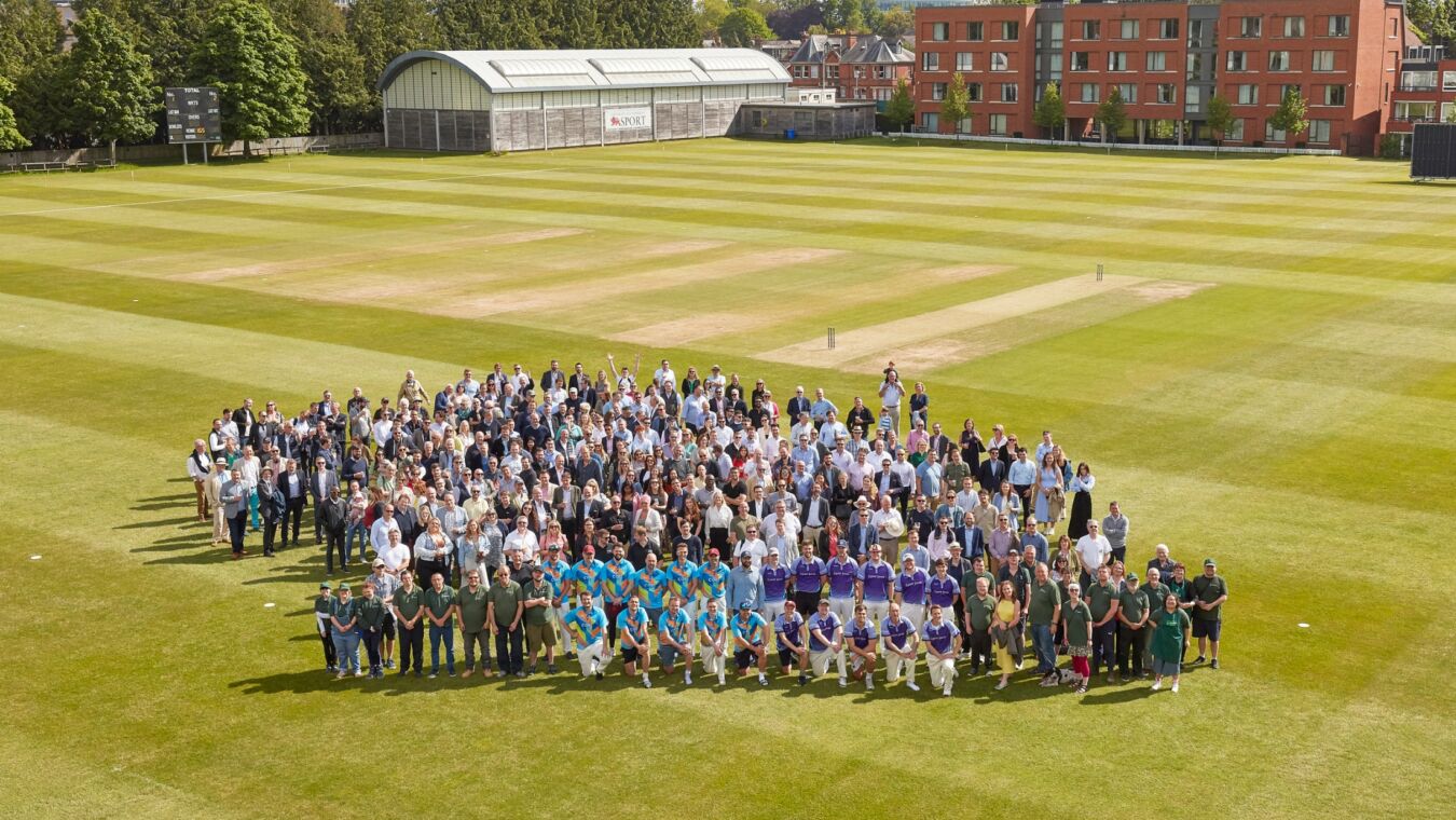 A large group gathers on a cricket field for a photo, surrounded by a scoreboard, pavilion, and residential buildings on a sunny day. The scoreboard displays "HOME 0" and "VISIT 0."