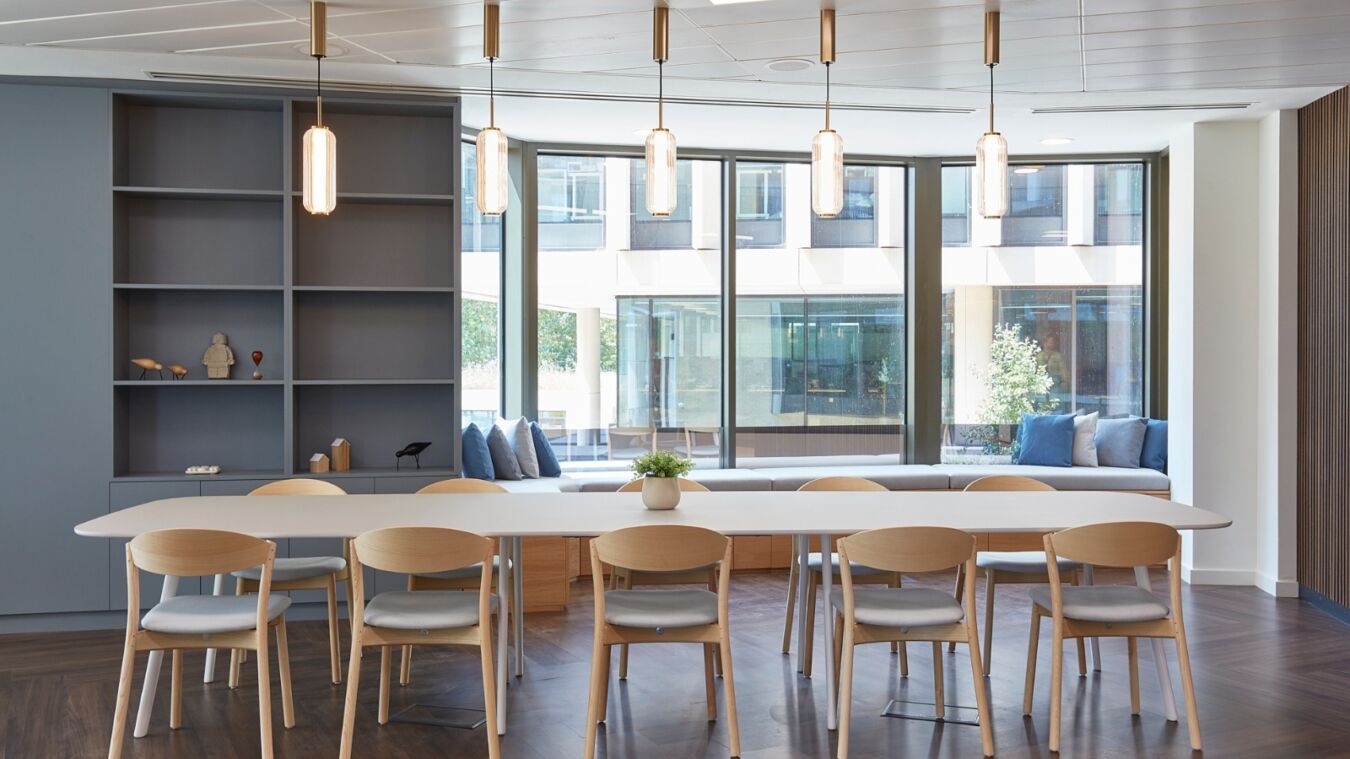 A modern dining area features a long table surrounded by wooden chairs, beneath pendant lights, near large windows. A blue cushioned bench and decorative shelves enhance the minimalist space.