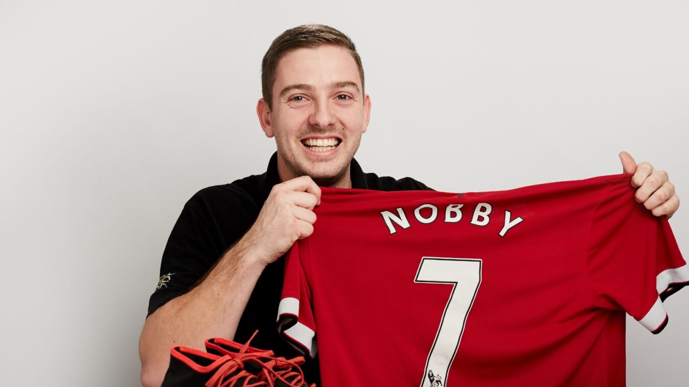 A person smiling, holds up a red football jersey with "NOBBY" and number 7, against a plain background. Soccer shoe with red laces visible in foreground.