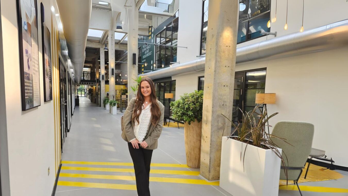 A woman stands smiling in a modern office hallway, surrounded by plants and industrial decor. A sign on the wall reads, "We nurture the low hums to make thunderous successes."