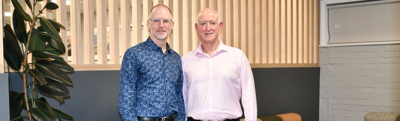 Two men stand smiling in an office with modern décor, featuring a large plant, wooden slat wall, colorful seating, and small tables.