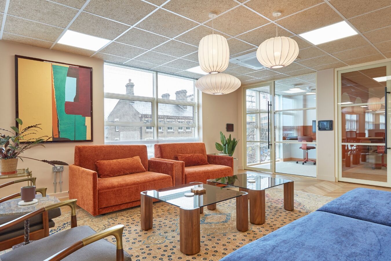 Stylish office lounge with orange armchairs, glass coffee tables, pendant lights, and large windows overlooking rooftops.
