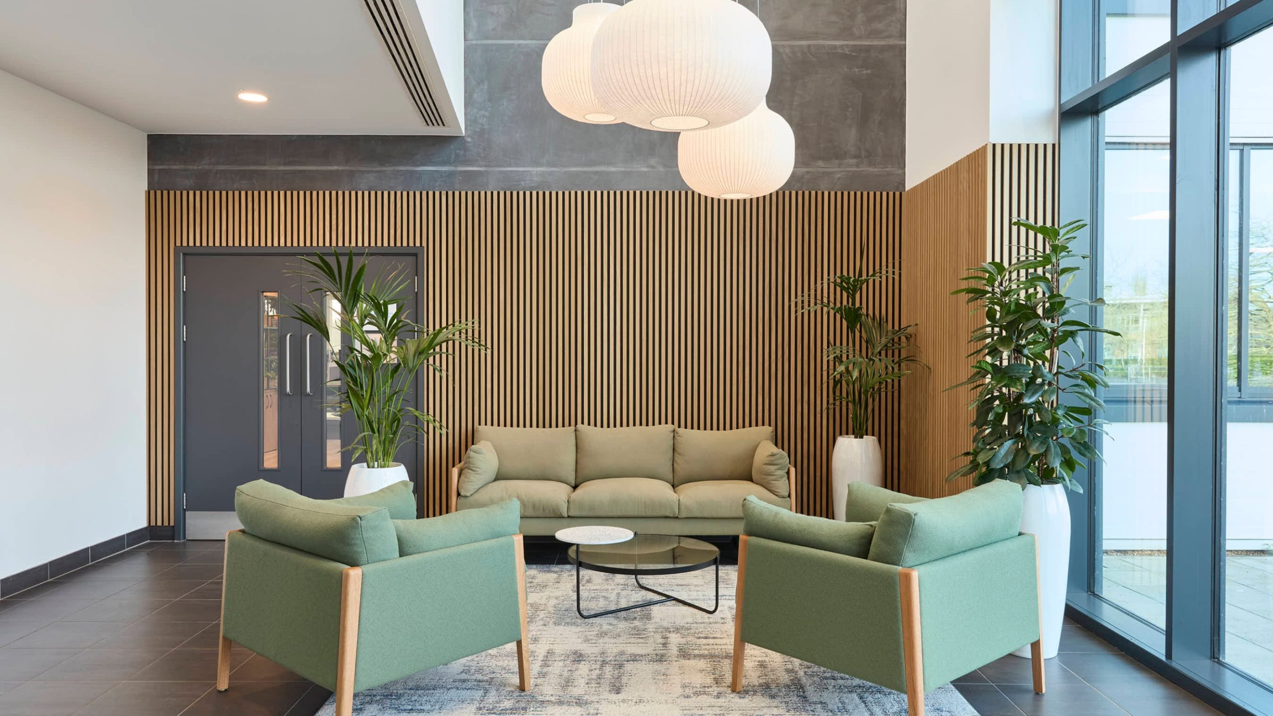 Modern lobby with a cozy feel; features green armchairs, a matching sofa, round glass coffee table, large plants, wood panel walls, and soft lighting