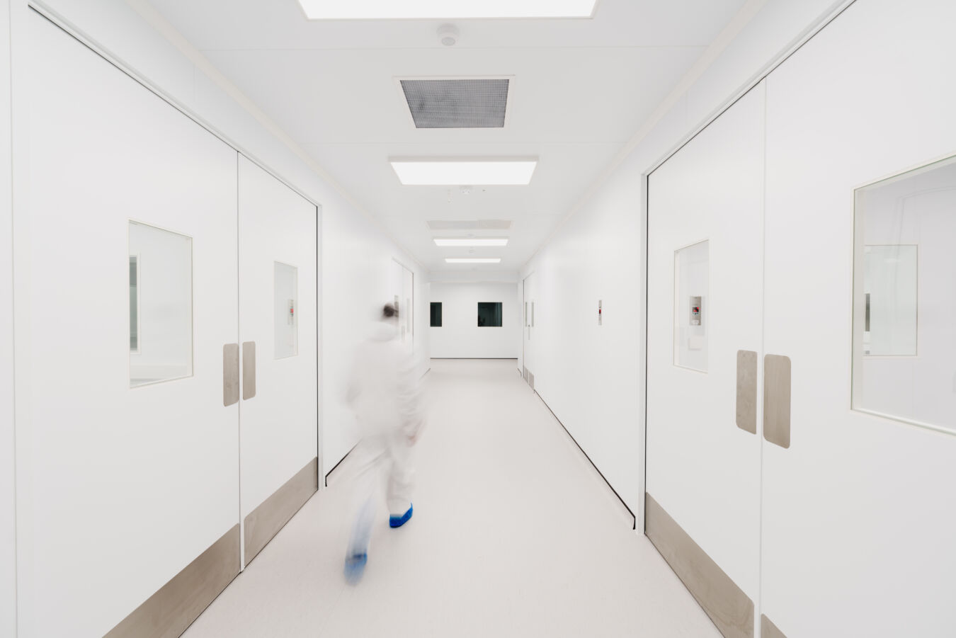 Person in protective clothing walking down a pristine, white corridor with large doors, bright lighting, and a sterile environment, suggesting a laboratory or cleanroom setting.