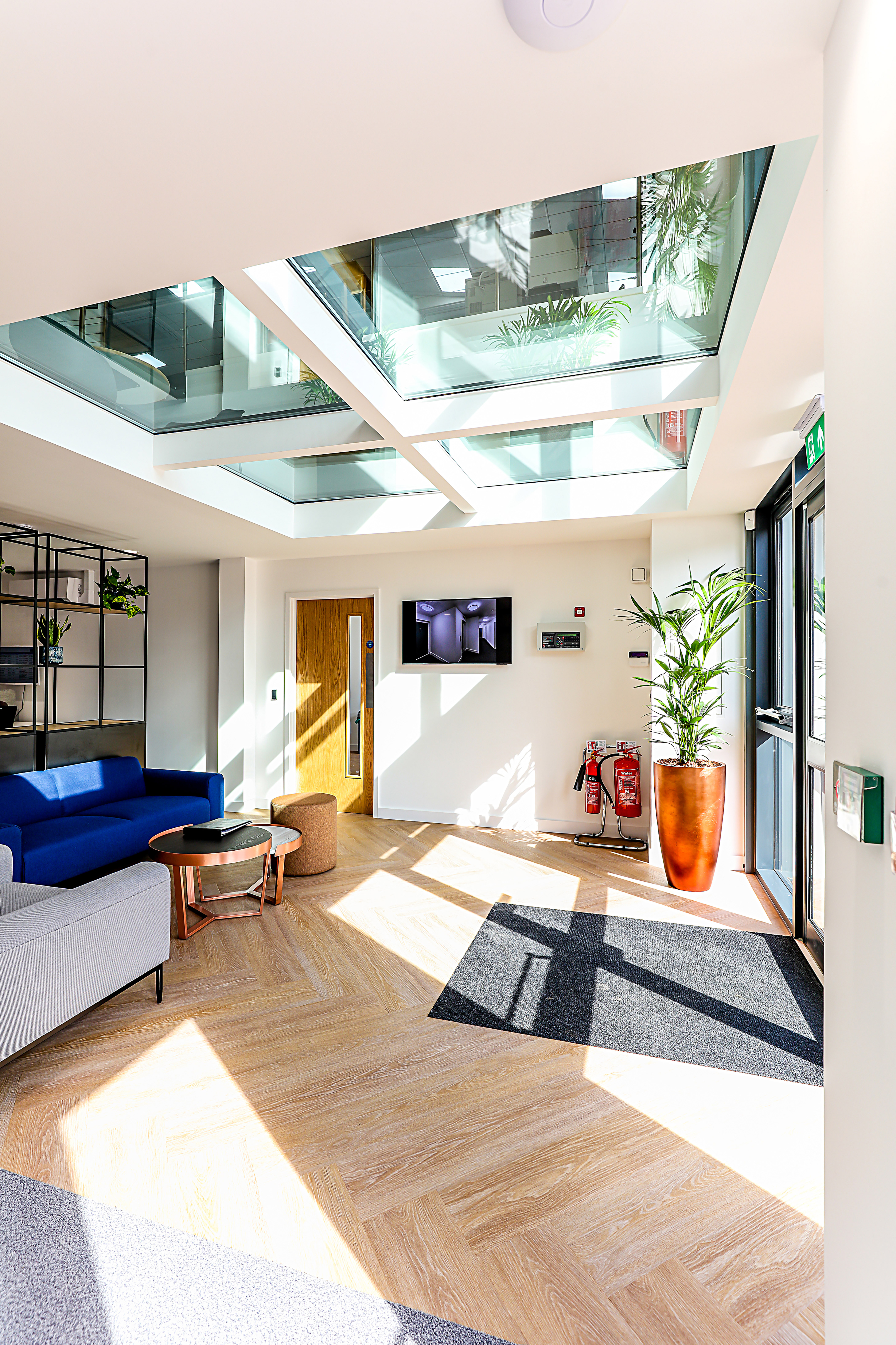Modern lobby with blue and gray sofas and a wooden table under natural light from a glass ceiling. The room has plants, fire extinguishers, and a wall-mounted screen displaying an interior scene.