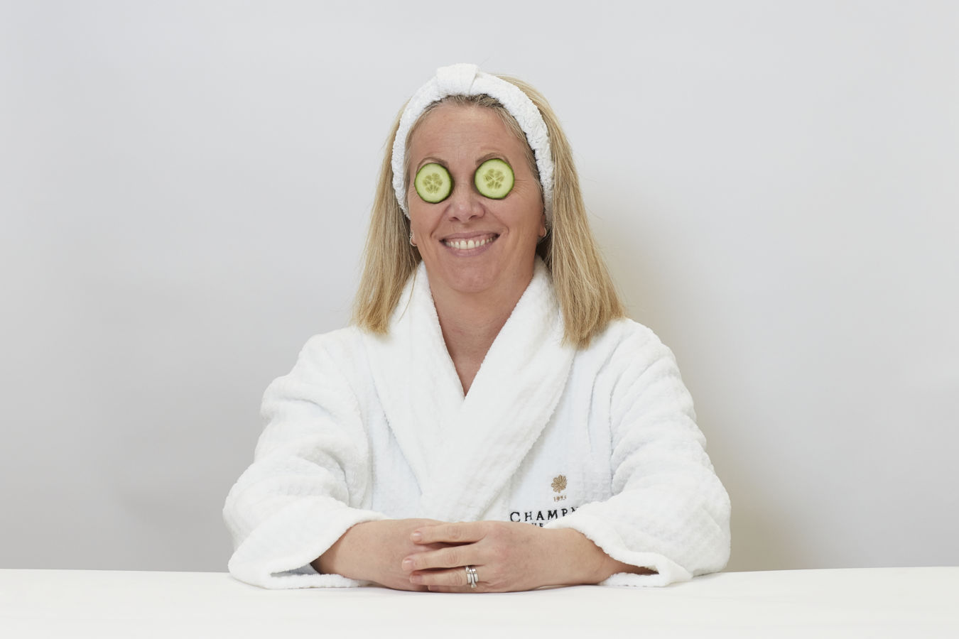 A person wearing a white robe with "CHAMPNEYS" logo and "1925" text, sits smiling with cucumber slices over their eyes, against a plain white background.