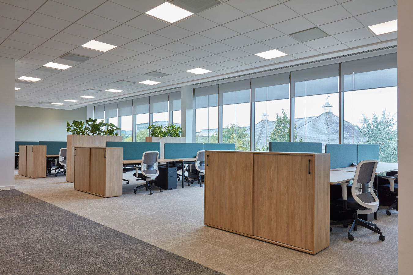 Modern office space with a row of workstations, ergonomic chairs, and wooden storage units. Large windows provide natural light, creating a bright, open atmosphere