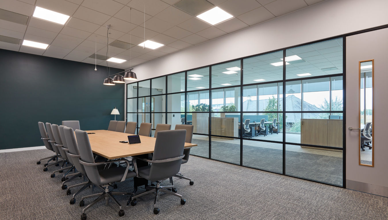 Modern conference room with a long wooden table, twelve gray chairs, and pendant lights. Glass wall reveals another office area with natural light