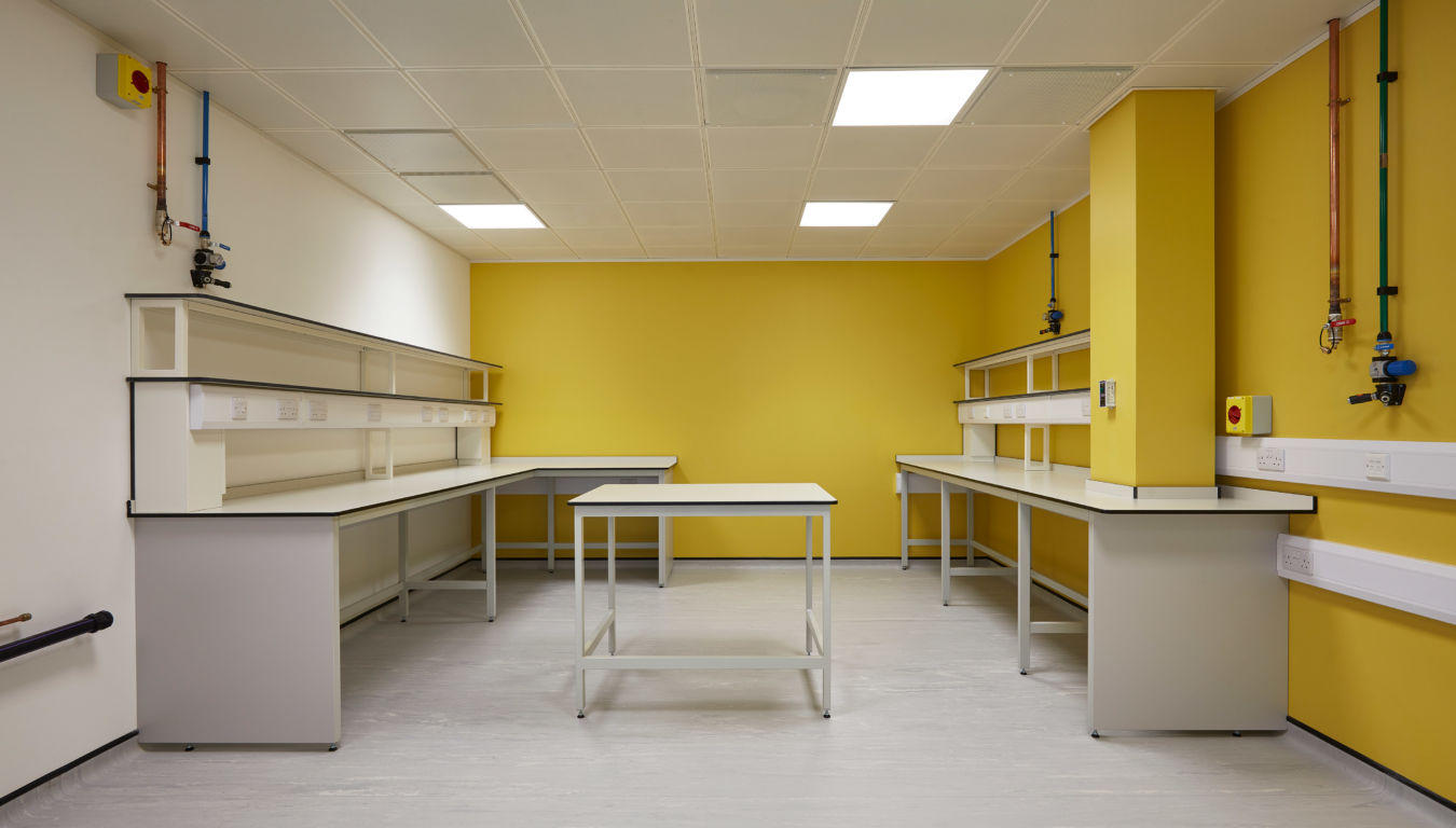A lab room features white tables with shelves against bright yellow walls, illuminated by ceiling lights. Various pipes and electrical outlets line the wall, suggesting a technical environment.