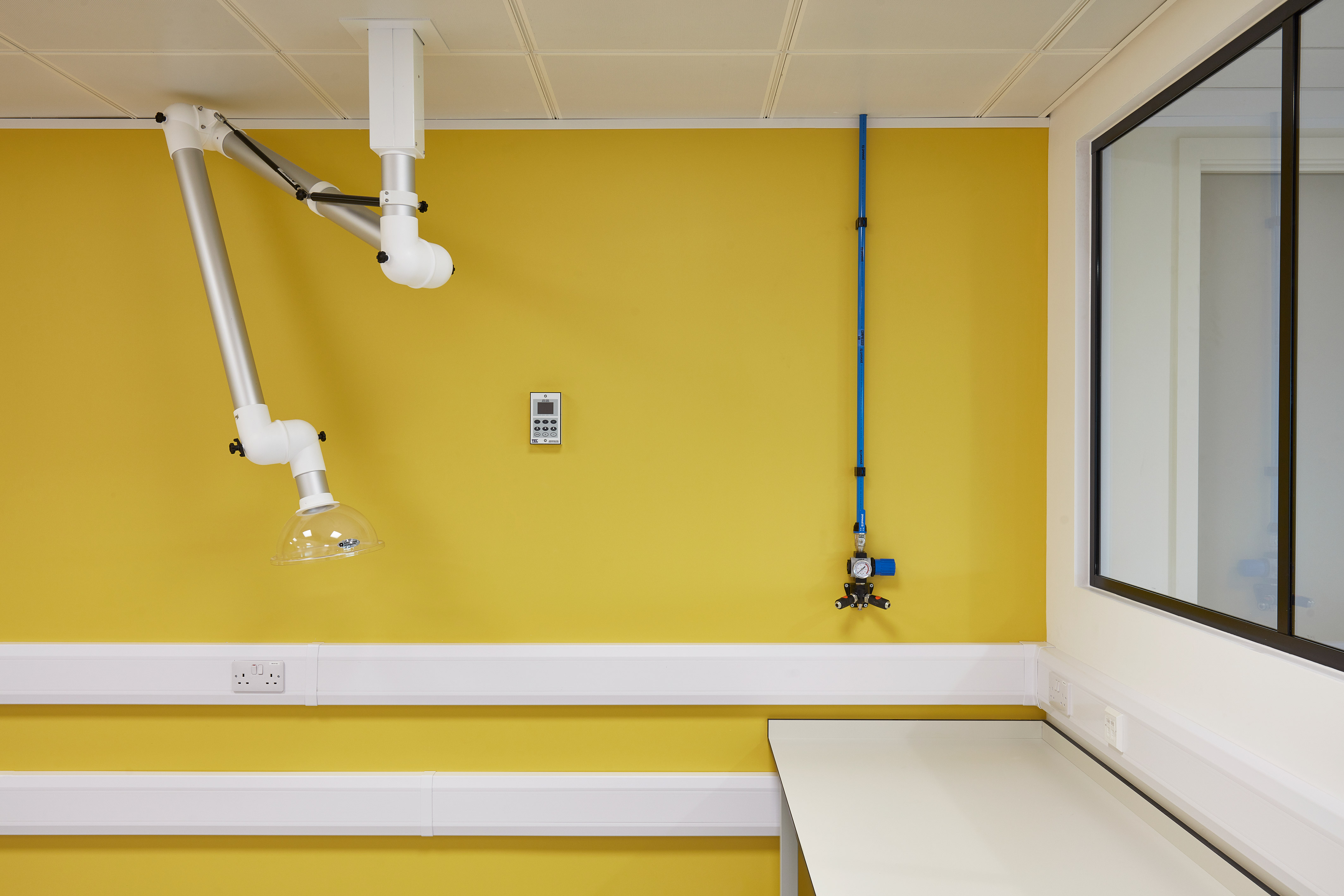An articulated arm with a transparent dome extends from the ceiling in a yellow-walled laboratory, near a digital control panel and a blue pipe system beside a window.