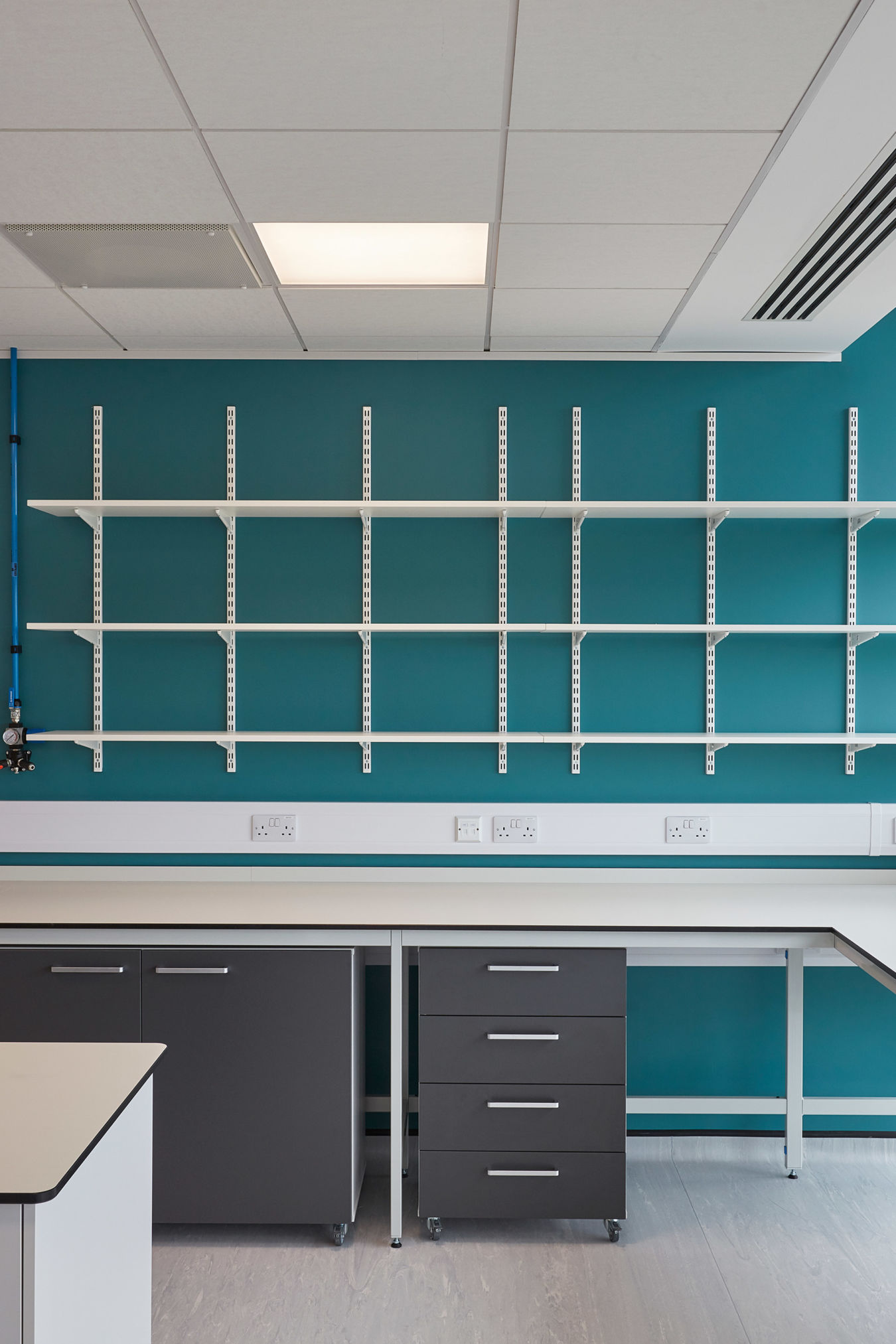 White wall-mounted shelves arranged in three horizontal rows on a teal-colored wall, above grey cabinets and drawers in a quiet, modern office space, with electrical outlets beneath.