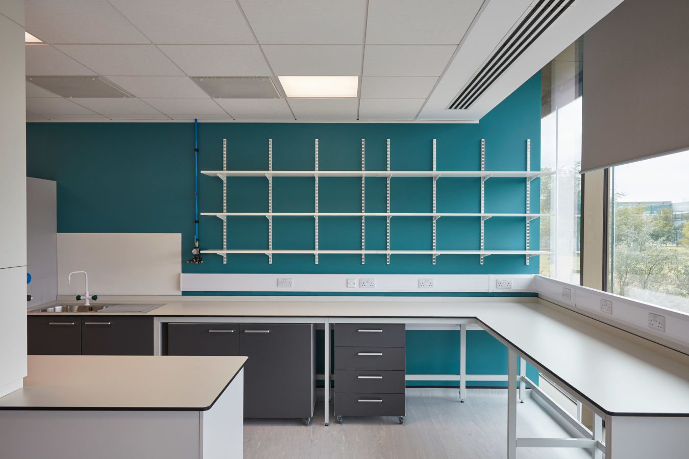 White wall-mounted shelves arranged in three horizontal rows on a teal-colored wall, above grey cabinets and drawers in a quiet, modern office space, with electrical outlets beneath.
