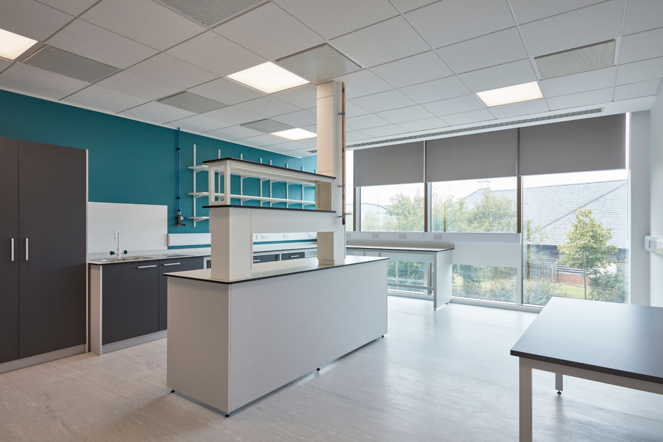 Laboratory countertops stand idle in a modern science lab, with shelves above and large windows providing natural light from outside greenery.