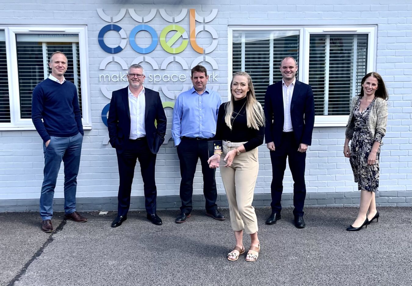 Six people standing in front of a white brick building featuring the "coel makes space work" logo; one person holds a transparent plaque, suggesting an award presentation.