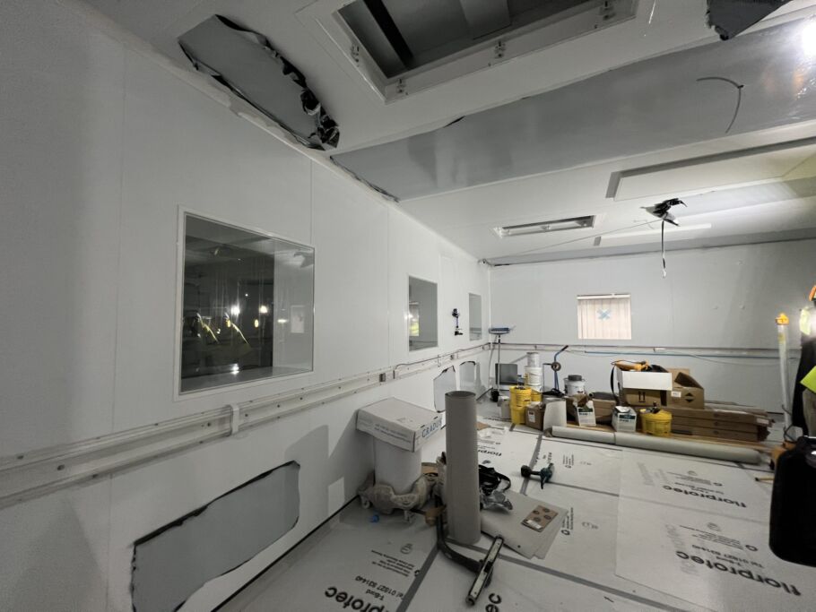 Interior of a cleanroom or laboratory under construction, with white walls, ceiling cutouts, observation windows, and equipment and materials scattered across the floor