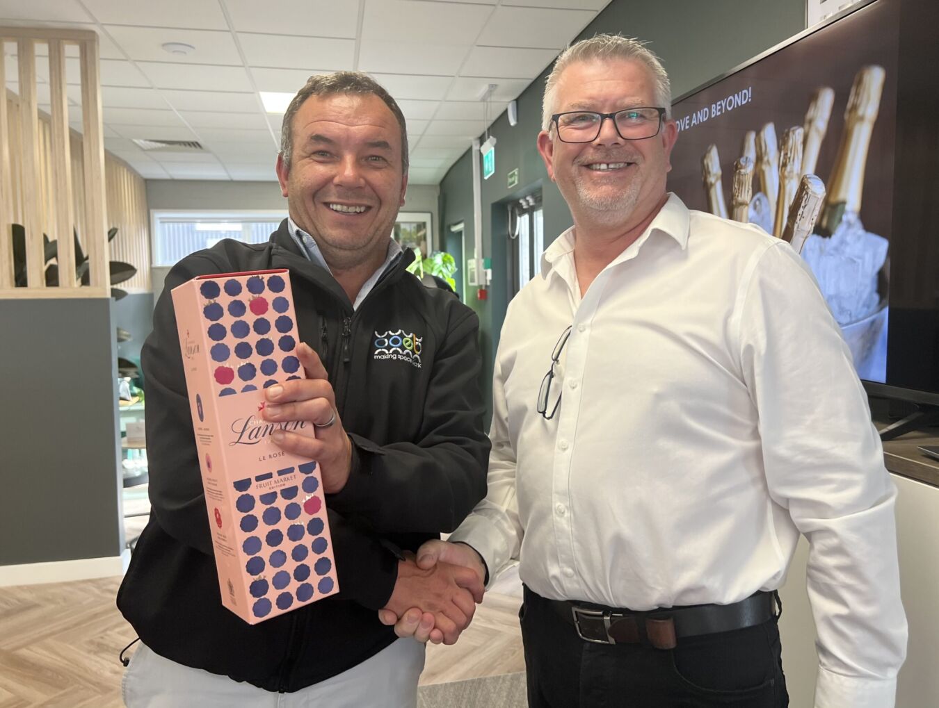 Two men smiling, one holding a pink box labeled "Lamura Le Rosé" with purple dots, shaking hands in a modern office with a large screen in the background.