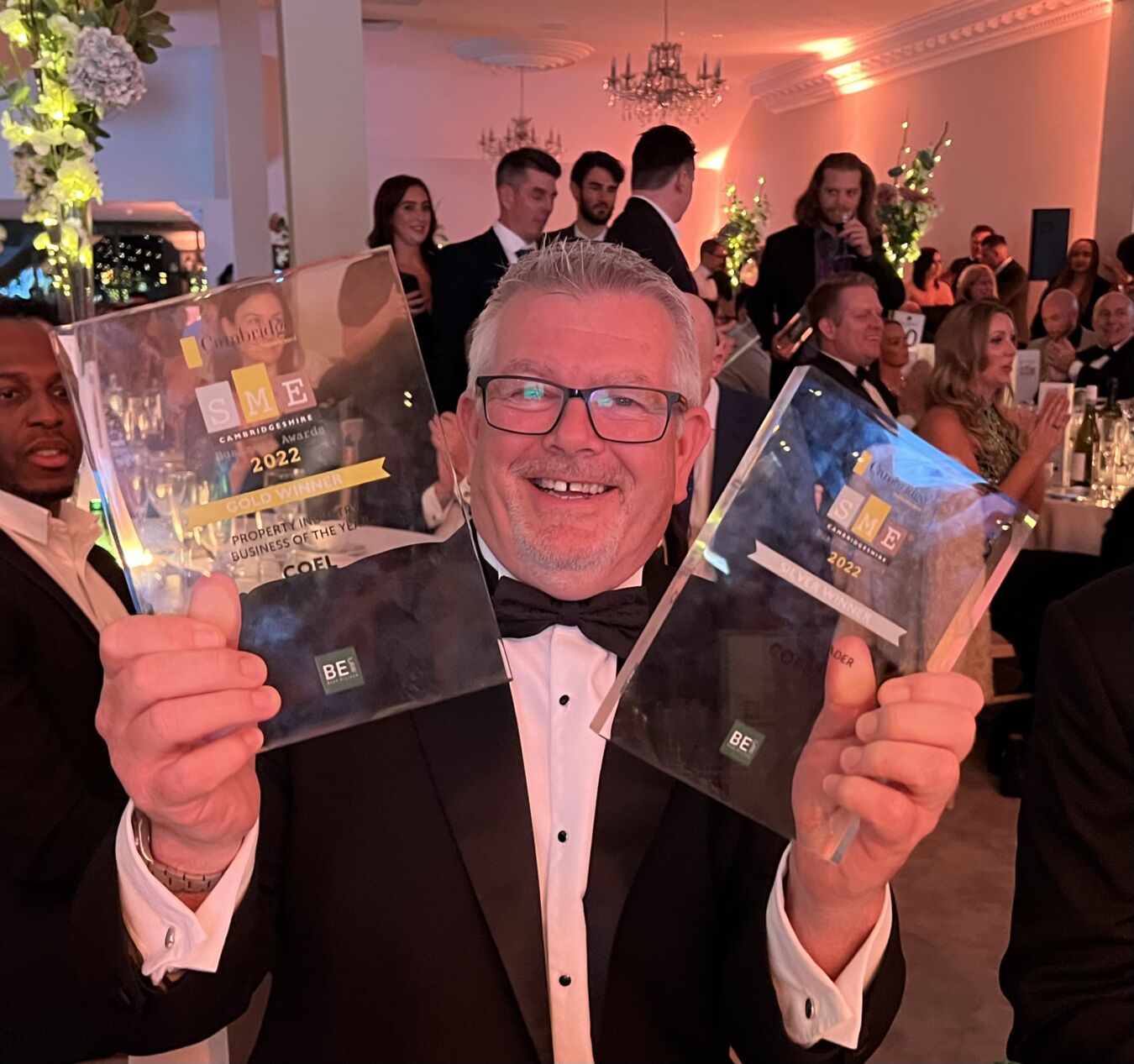 A man in a tuxedo holds two SME Cambridgeshire Awards from 2022, labeled "Gold Winner" for Property and Business of the Year, in a formal event with seated guests.
