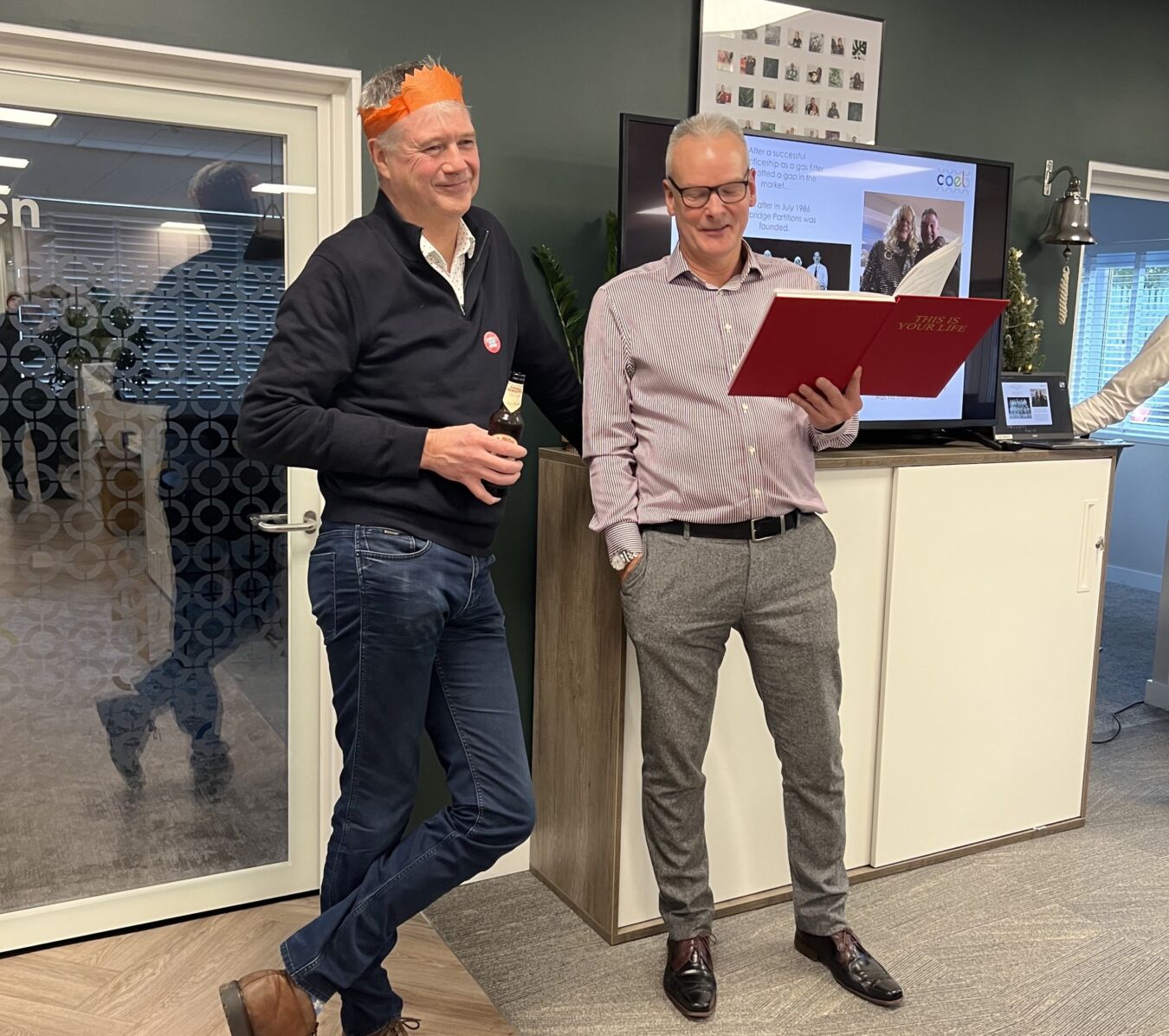 Two men stand in an office; one in a black sweater and orange headband holds a drink, while the other reads from a red folder labeled "This is your life." A screen displays part of a presentation.