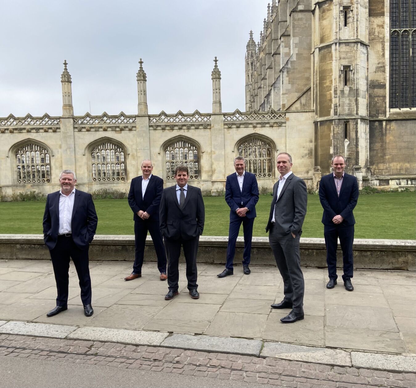 Six men in suits stand on a stone path, posing in front of a historic building with ornate windows and spires, surrounded by green lawn and overcast sky.